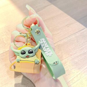 Op de afbeelding: Een groene en gele sleutelhanger met een stripfiguur. De sleutelhanger heeft een gouden ring en een groene band met de tekst "RAW RAW". Het personage heeft grote zwarte ogen en een kleine glimlach.