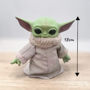 K&ouml;nnte beinhalten: Eine gr&uuml;ne Figur einer Figur mit gro&szlig;en schwarzen Augen und gro&szlig;en Ohren, die einen beigen Mantel mit flauschigem Kragen tr&auml;gt. Die Figur ist 18 cm gro&szlig;.