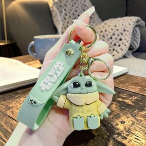 Op de afbeelding: Een groene en gele Baby Yoda sleutelhanger met een goudkleurige sleutelring. De sleutelhanger heeft een groene band met de tekst "STAR WARS". Het Baby Yoda figuur draagt een gele jurk.