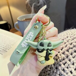 Op de afbeelding: Een groene en gele Grogu sleutelhanger met een goudkleurige sleutelring. De sleutelhanger heeft een groene band met de tekst "STAR WARS" en een Grogu gezicht. Het Grogu figuur houdt een zwarte sjaal vast.