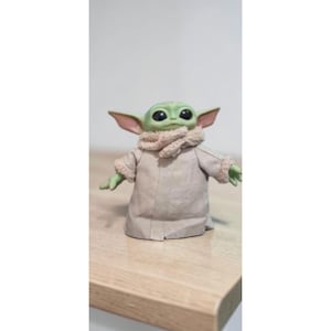 K&ouml;nnte beinhalten: Eine gr&uuml;ne Grogu-Figur, auch bekannt als Baby Yoda, steht auf einer Holzoberfl&auml;che. Die Figur hat gro&szlig;e schwarze Augen, spitze Ohren und tr&auml;gt eine beige Robe mit einem beigen Schal. Die Figur ist ein Charakter aus dem Star Wars-Franchise.