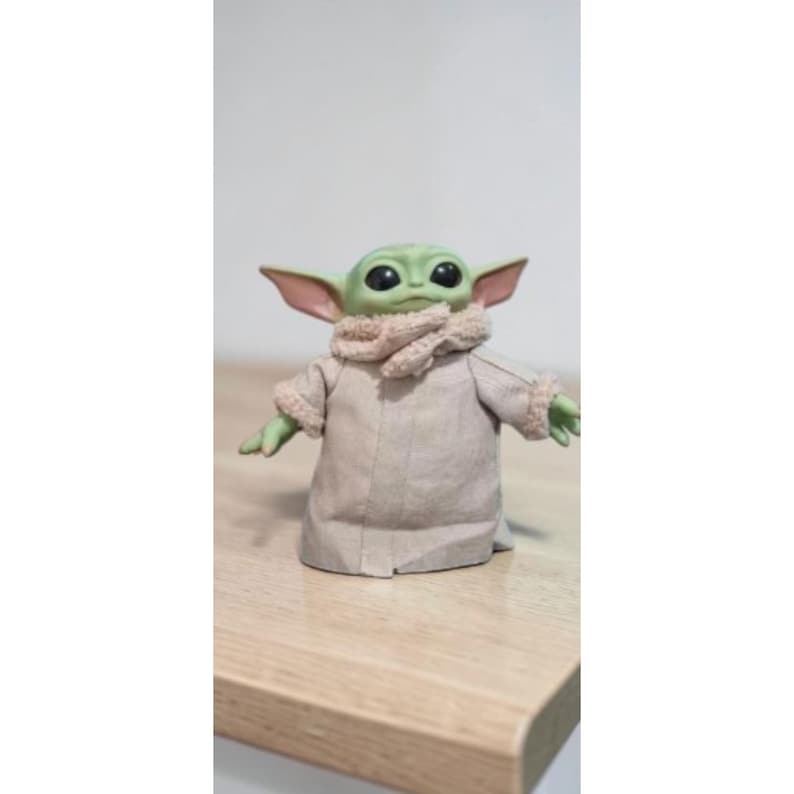 K&ouml;nnte beinhalten: Eine gr&uuml;ne Grogu-Figur, auch bekannt als Baby Yoda, steht auf einer Holzoberfl&auml;che. Die Figur hat gro&szlig;e schwarze Augen, spitze Ohren und tr&auml;gt eine beige Robe mit einem beigen Schal. Die Figur ist ein Charakter aus dem Star Wars-Franchise.