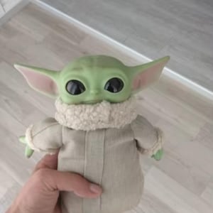 K&ouml;nnte beinhalten: Eine gr&uuml;n geh&auml;utete Grogu-Puppe mit gro&szlig;en schwarzen Augen und spitzen Ohren. Die Puppe tr&auml;gt einen beigefarbenen Umhang mit einem flauschigen, cremefarbenen Kragen. Die Arme der Puppe sind ausgestreckt und sie hat kleine gr&uuml;ne H&auml;nde.