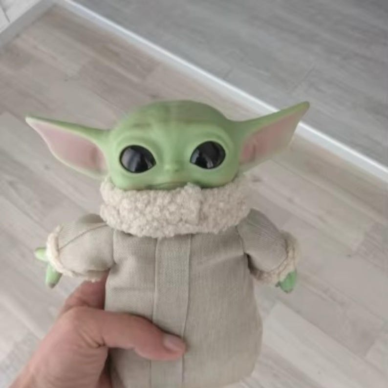 K&ouml;nnte beinhalten: Eine gr&uuml;n geh&auml;utete Grogu-Puppe mit gro&szlig;en schwarzen Augen und spitzen Ohren. Die Puppe tr&auml;gt einen beigefarbenen Umhang mit einem flauschigen, cremefarbenen Kragen. Die Arme der Puppe sind ausgestreckt und sie hat kleine gr&uuml;ne H&auml;nde.