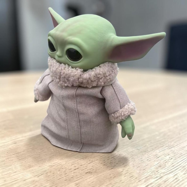 K&ouml;nnte beinhalten: Eine gr&uuml;n geh&auml;utete Figur mit gro&szlig;en Augen und Ohren, bekleidet mit einem hellrosa Mantel mit flauschigem Kragen und Manschetten. Die Figur ist ein Charakter aus einem beliebten Science-Fiction-Franchise.