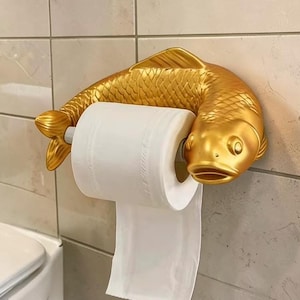 Puede incluir: Un portarrollos de papel higiénico de color dorado con forma de pez. El cuerpo del pez se arquea sobre la parte superior de un rollo de papel higiénico blanco estándar, con la cabeza a la derecha y la cola a la izquierda, sujetando el rollo.