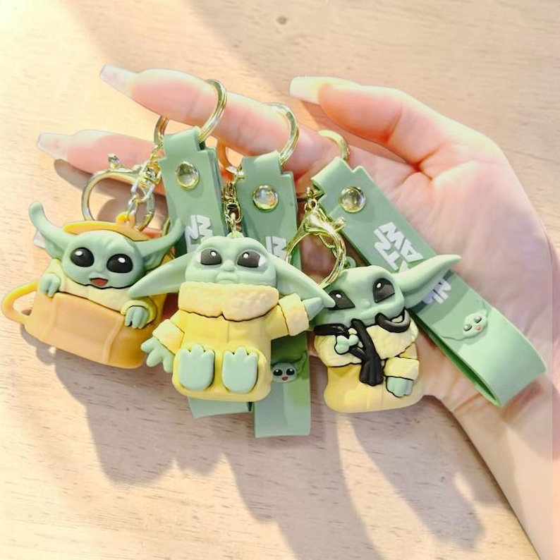Op de afbeelding: Vier sleutelhangers met een groen alien-personage met grote ogen en puntige oren. Het personage draagt een gele outfit. Elke sleutelhanger heeft een groene band en een goudkleurige ring.