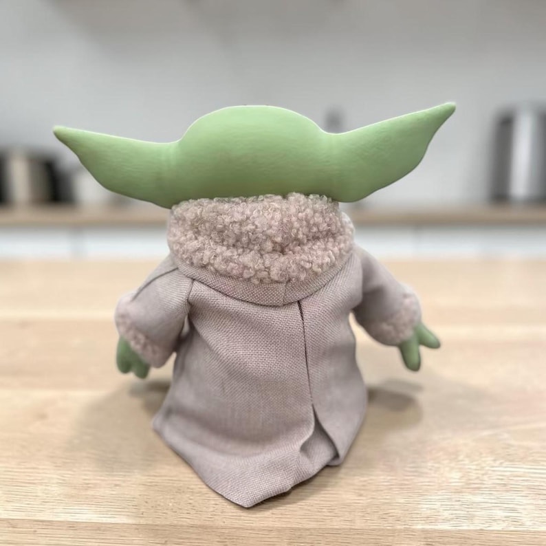 K&ouml;nnte beinhalten: Pl&uuml;schpuppe einer Figur mit gro&szlig;en gr&uuml;nen Ohren, die einen beigen Mantel mit flauschigem Kragen tr&auml;gt. Die H&auml;nde und F&uuml;&szlig;e der Puppe sind gr&uuml;n. Die Puppe ist eine beliebte Figur aus einem Science-Fiction-Franchise.