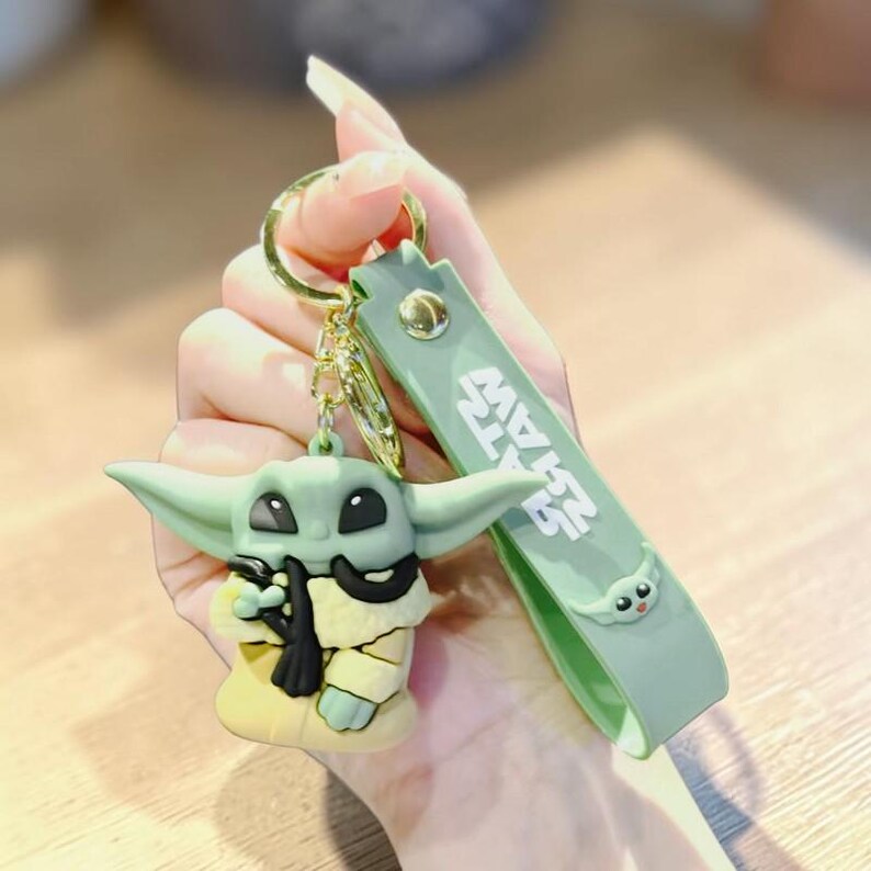 Op de afbeelding: Een groene en gele sleutelhanger met een stripfiguur met grote oren en zwarte ogen. De sleutelhanger heeft een gouden ring en een groene band met de tekst "STAR WARS".