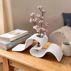 Puede incluir: Un florero de cerámica blanca con una forma ondulada continua que presenta una única abertura circular en la parte superior. El florero sostiene una rama delicada con pequeñas flores de color rosa claro, creando un contraste con la superficie blanca mate.