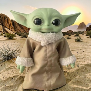 K&ouml;nnte beinhalten: Puppe einer Figur mit gro&szlig;en schwarzen Augen, gr&uuml;ner Haut und gro&szlig;en Ohren. Die Puppe tr&auml;gt eine beige Robe mit wei&szlig;em, flauschigem Besatz. Der Hintergrund zeigt eine W&uuml;stenlandschaft mit Sonnenuntergang.