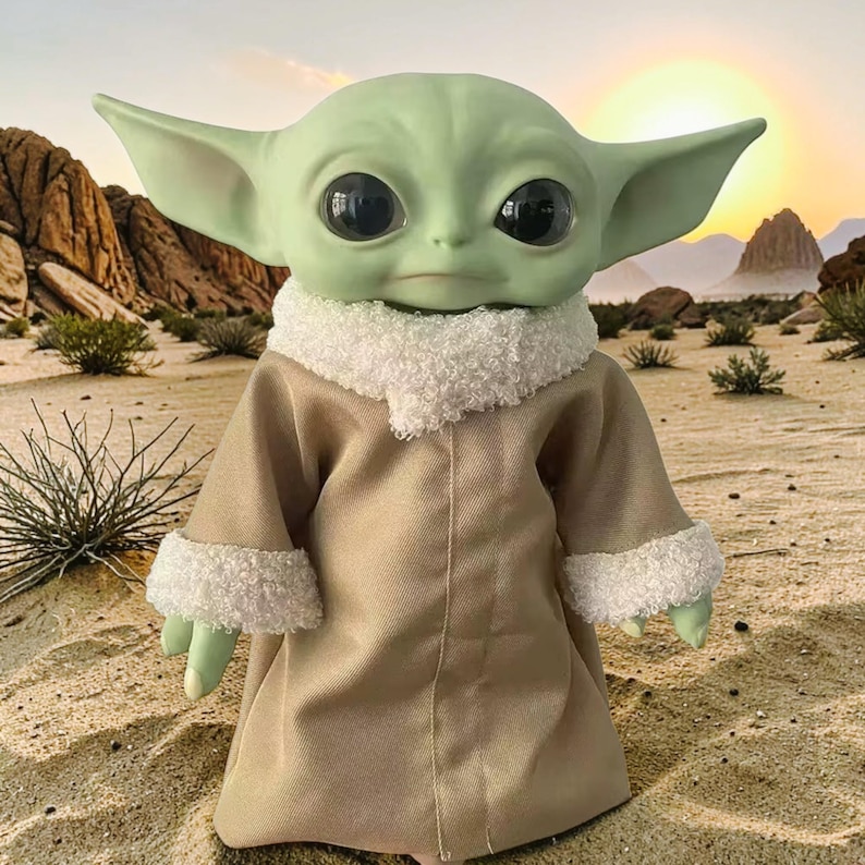 K&ouml;nnte beinhalten: Puppe einer Figur mit gro&szlig;en schwarzen Augen, gr&uuml;ner Haut und gro&szlig;en Ohren. Die Puppe tr&auml;gt eine beige Robe mit wei&szlig;em, flauschigem Besatz. Der Hintergrund zeigt eine W&uuml;stenlandschaft mit Sonnenuntergang.