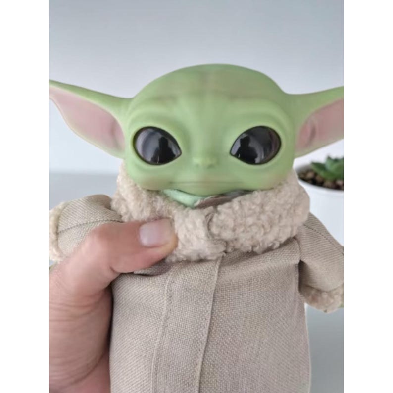 K&ouml;nnte beinhalten: Eine Puppe mit gr&uuml;ner Haut, gro&szlig;en schwarzen Augen und spitzen Ohren. Die Puppe tr&auml;gt einen beigefarbenen Mantel mit einem flauschigen, cremefarbenen Kragen. Die Puppe ist eine Figur aus einem beliebten Science-Fiction-Franchise.