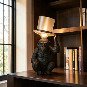 May include: A table lamp shaped like a seated dark brown chimpanzee holding a translucent white top hat over its head. The hat serves as a lampshade, diffusing light from a bulb positioned between the primate's head and the hat's brim.