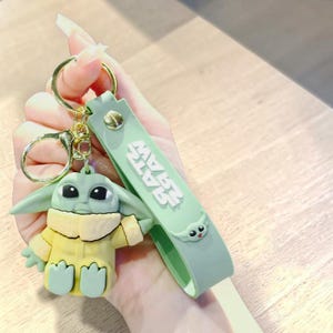 Op de afbeelding: Een groene en gele Baby Yoda sleutelhanger met een goudkleurige sleutelring en een groene band. Op de band staat de tekst "STAR WARS". De sleutelhanger is een schattige, cartoonachtige weergave van het personage.