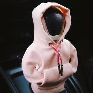 Puede incluir: Una funda para palanca de cambios con forma de sudadera con capucha rosa claro, con un bolsillo delantero y un cordón rojo. Diseñada para encajar sobre un pomo de palanca de cambios negro, añadiendo un toque único y elegante al interior de un vehículo.