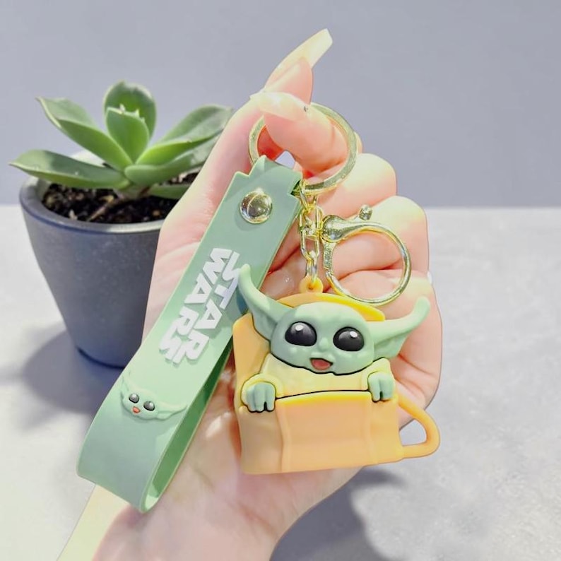 Op de afbeelding: Een groene en gele Star Wars sleutelhanger met een Grogu-personage. De sleutelhanger heeft een groene band met de woorden "STAR WARS" erop gedrukt. Het Grogu-figuur zit in een gele beker en heeft grote zwarte ogen.