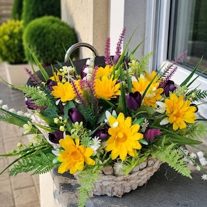 Op de afbeelding: Een bloemstuk in een gevlochten mand met felgele madeliefjes, donkerpaarse tulpen, witte lelietjes-van-dalen en groene varenbladeren. Het arrangement bevat hoge paarse aren en divers groen blad, wat zorgt voor een dicht en kleurrijk geheel.