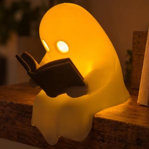 May include: A glowing yellow ghost-shaped lamp with two circular eye cutouts, holding a small dark-colored open book. The lamp is positioned on the edge of a wooden surface, emitting a warm light from within its smooth, rounded form.
