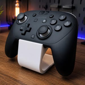 Pode incluir: Um controle preto Nintendo Switch Pro em um suporte branco. O controle possui um direcional, manípulos analógicos e botões de ação. O suporte é curvo e projetado para segurar o controle na vertical.