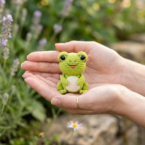May include: A small, light green crocheted frog figurine with a round, cream-colored belly, large black plastic eyes, and a tiny pink stitched mouth. The frog is crafted with a textured yarn stitch pattern and has a rounded head and body.