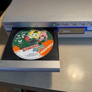 May include: A silver Sony DVD player with an open tray displaying a Nickelodeon Christmas DVD. The disc features SpongeBob SquarePants and Patrick Star. The word "Christmas" is visible on the disc.