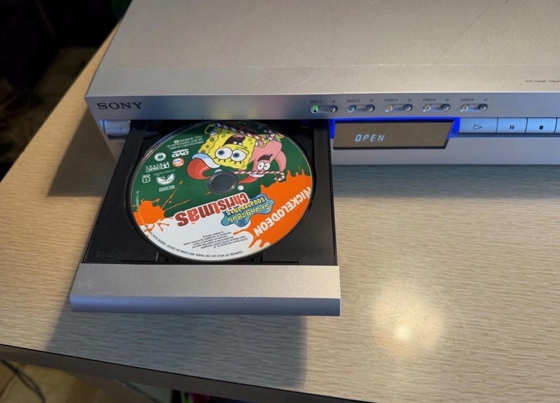 May include: A silver Sony DVD player with an open tray displaying a Nickelodeon Christmas DVD. The disc features SpongeBob SquarePants and Patrick Star. The word "Christmas" is visible on the disc.
