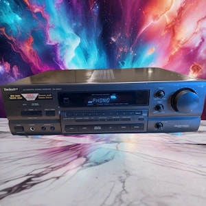 Puede incluir: Un receptor estéreo de control AV Technics modelo SA-GX67 de color gris oscuro con una pantalla digital que muestra PHONO, múltiples botones para la selección de entrada, un dial de volumen grande y perillas para ajustes de agudos, graves y balance.