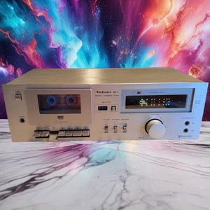 May include: A silver Technics M11 stereo cassette deck featuring a front-loading tape compartment, a digital fluorescent meter display, a large volume control knob, multiple push-button controls, and input jacks for microphones and headphones.