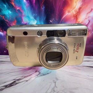 May include: A silver Canon Sure Shot Z180u point-and-shoot camera featuring a Canon Zoom Lens 38-180mm. The front of the camera displays the brand name Canon, the model name Sure Shot Z180u, and a built-in flash unit positioned on the top right side.