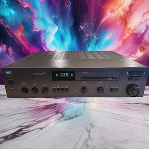 May include: A dark grey NAD 7105PE stereo receiver featuring a digital display showing 550, multiple control knobs for bass, treble, and balance, a headphone jack, and various push-button inputs on the front panel for audio equipment control.