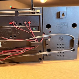 May include: A close-up of a vintage black and gray Sony boombox. The back of the boombox is visible, showing the speaker system and various connection ports. Red and black wires are connected to the boombox. The speaker system is labeled "SONY SPEAKER SYSTEM LEFT".