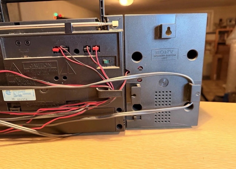 May include: A close-up of a vintage black and gray Sony boombox. The back of the boombox is visible, showing the speaker system and various connection ports. Red and black wires are connected to the boombox. The speaker system is labeled "SONY SPEAKER SYSTEM LEFT".