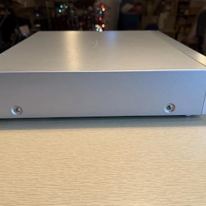 May include: A silver-colored electronic device with a rectangular shape. The device has a smooth, reflective surface and visible screws on the bottom. The item is placed on a light-colored wooden surface.