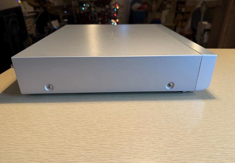 May include: A silver-colored electronic device with a rectangular shape. The device has a smooth, reflective surface and visible screws on the bottom. The item is placed on a light-colored wooden surface.