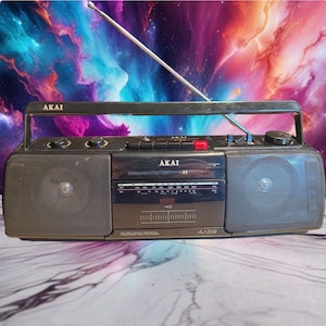 May include: A black Akai AJ-208 portable radio cassette player featuring a top handle, a long silver telescopic antenna, two side speakers, and a central control panel with a radio frequency display, volume knobs, and playback buttons.