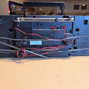 May include: A black Sony boombox with a handle, antenna, and various ports and wires visible on the back. The boombox is sitting on a light brown surface. The back of the boombox has the word "CAUTION" printed on it.