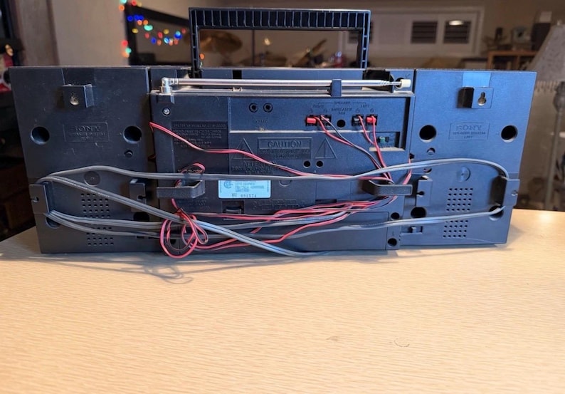 May include: A black Sony boombox with a handle, antenna, and various ports and wires visible on the back. The boombox is sitting on a light brown surface. The back of the boombox has the word "CAUTION" printed on it.