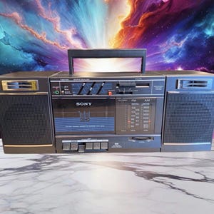 May include: A vintage Sony boombox with a black and silver finish. The boombox features a cassette player, radio tuner, and graphic equalizer. The handle is black, and the speakers are covered with a black mesh. The background is a colorful nebula.