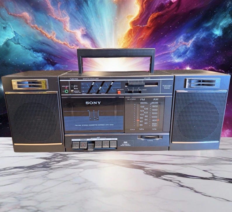 May include: A vintage Sony boombox with a black and silver finish. The boombox features a cassette player, radio tuner, and graphic equalizer. The handle is black, and the speakers are covered with a black mesh. The background is a colorful nebula.