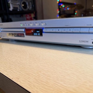 May include: Silver Sony DVD player with a digital display showing "000 18". The device has several buttons and a headphone jack. The text "S-master" is visible on the front. The player is on a light wood-grain surface.
