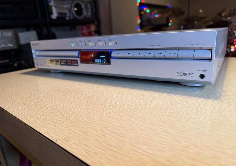 May include: Silver Sony DVD player with a digital display showing "000 18". The device has several buttons and a headphone jack. The text "S-master" is visible on the front. The player is on a light wood-grain surface.