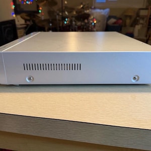 May include: A silver-colored electronic device with a rectangular shape, featuring a series of ventilation slots and two visible screws. The device is placed on a light-colored wooden surface, suggesting a home entertainment or audio component.