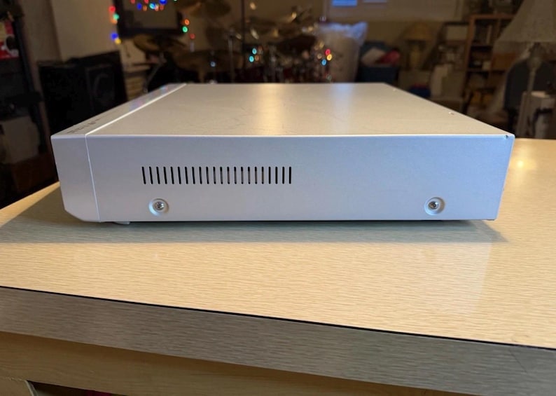 May include: A silver-colored electronic device with a rectangular shape, featuring a series of ventilation slots and two visible screws. The device is placed on a light-colored wooden surface, suggesting a home entertainment or audio component.
