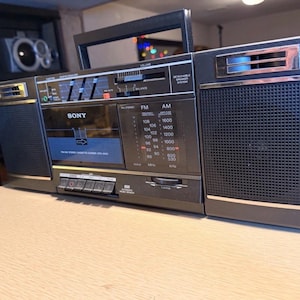 May include: A vintage black Sony boombox with a cassette player, AM/FM radio, and a handle. The boombox has a silver trim, a speaker on each side, and various dials and buttons. The radio dial displays FM and AM frequencies.