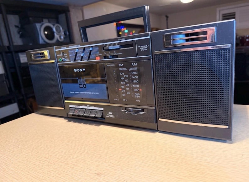 May include: A vintage black Sony boombox with a cassette player, AM/FM radio, and a handle. The boombox has a silver trim, a speaker on each side, and various dials and buttons. The radio dial displays FM and AM frequencies.