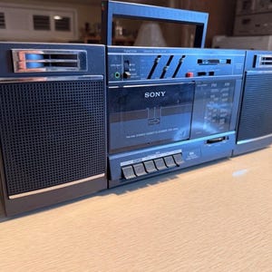 May include: A vintage Sony boombox in shades of blue and black, featuring a cassette player and radio. The boombox has a handle for portability and a speaker on each side. The Sony logo is visible on the front.