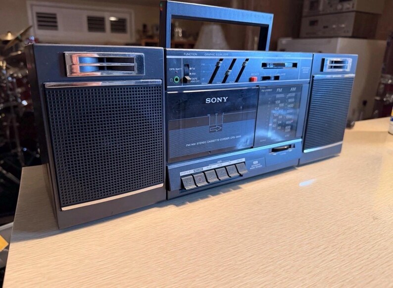 May include: A vintage Sony boombox in shades of blue and black, featuring a cassette player and radio. The boombox has a handle for portability and a speaker on each side. The Sony logo is visible on the front.