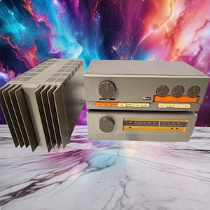 May include: A grey Quad audio system consisting of a power amplifier with vertical cooling fins, a preamplifier with multiple circular control knobs and orange input buttons, and a radio tuner with a horizontal orange frequency display panel.