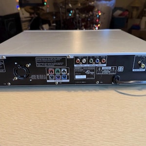 May include: Silver and black Sony DVD player with various input and output ports. The back panel features multiple connection options, including video, audio, and speaker ports. The device has a power cord and a fan for cooling.